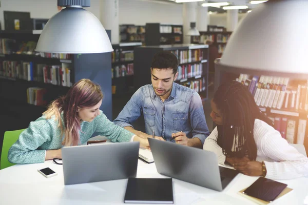 Crew of skilled male and female international students learning together preparing for examination in university library using modern technologies